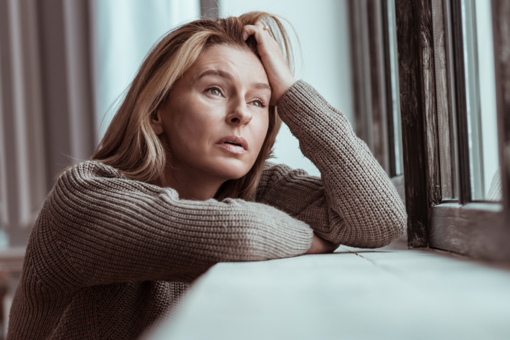 A woman sitting by a window, appearing contemplative and distressed, symbolizing the emotional toll of self-neglect